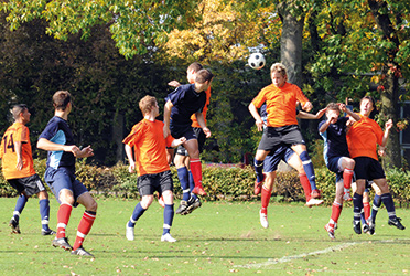 Das Bild zeigt zwei Mannschaften beim Fußballspiel. Einer der Spieler macht gerade einen Kopfball. (verweist auf: Kopf­verletzungen im Fußball und die kontroverse Diskussion um das Spielen von Kopf­bällen)